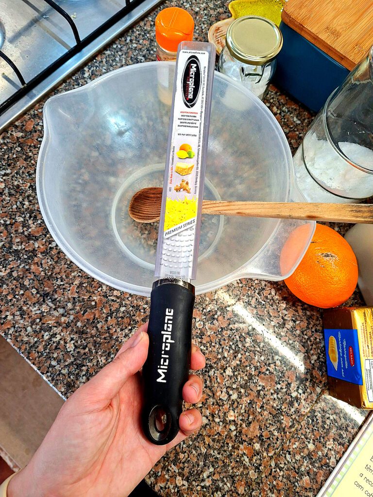 Hand holding Microplane Premium Series grater with black ergonomic handle, on granite counter with mixing bowl, orange and ingredients in the background