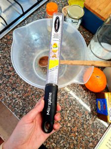 Mão a segurar ralador Microplane Premium Series com cabo ergonómico preto, sobre bancada de granito com tigela, laranja e ingredientes ao fundo