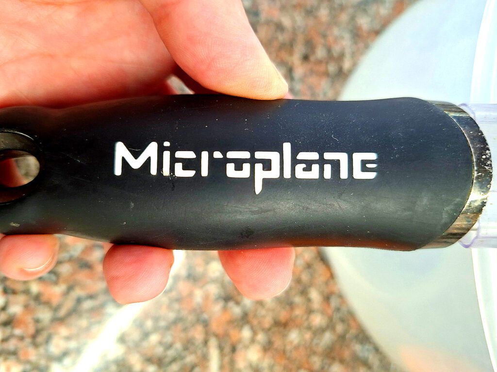 Close-up do cabo ergonómico preto do ralador Microplane com marca gravada em branco, segurado por mão adulta