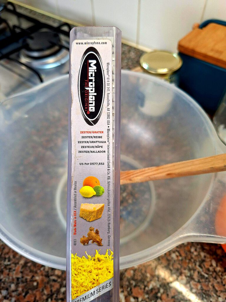 Embalagem do ralador Microplane Premium Series Zester Grater com informações do produto, imagens de limão, laranja, gengibre e queijo, com tigela de cozinha ao fundo