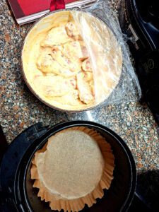 A bowl of marinated chicken next to an air fryer lined with parchment paper, in a home kitchen setting.