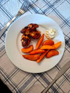 Prato com asinhas de frango assadas, batatas rústicas e molho cremoso, sobre toalha aos quadrados, com talheres ao lado.