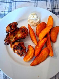 Prato com três asinhas de frango assadas, batatas douradas e molho branco, apresentado numa mesa com toalha aos quadrados.