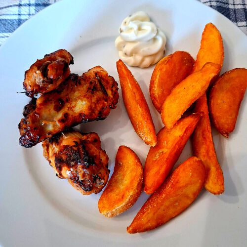 Prato com três asinhas de frango assadas, batatas douradas e molho branco, apresentado numa mesa com toalha aos quadrados.