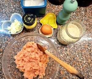 Ingredients for homemade chicken rolls laid out on a countertop: chopped chicken breasts, egg, onion, garlic, Dijon mustard, breadcrumbs, Flamengo cheese, and porchetta