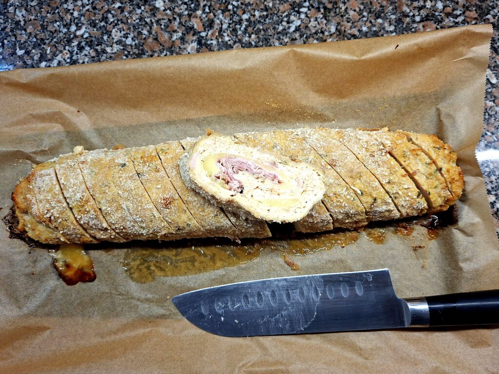 Rolo de frango caseiro fatiado sobre papel vegetal, com recheio de queijo e porchetta visível ao centro, acompanhado de faca de chef