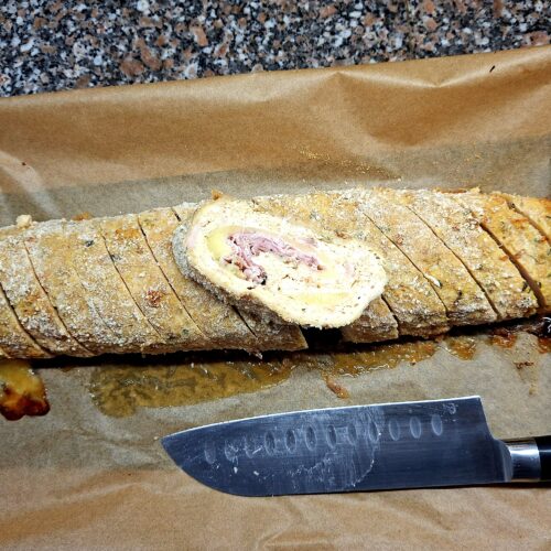 Rolo de frango caseiro fatiado sobre papel vegetal, com recheio de queijo e porchetta visível ao centro, acompanhado de faca de chef