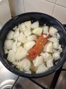 Dark blue pot on a gas stove with quartered onions and a whole chouriço sausage cooking in broth.