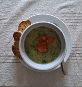White bowl with caldo verde soup, three slices of chouriço and a drizzle of olive oil at the centre, served alongside slices of cornbread on a plate, seen from above.