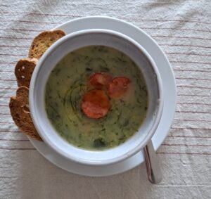 Tigela branca com caldo verde denso e rodelas de chouriço vista de cima, com quatro fatias de broa de milho ao lado, sobre toalha de linho às riscas.