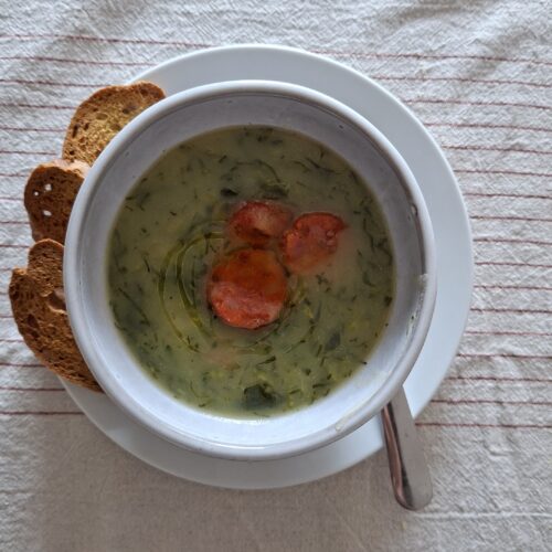 Tigela branca com caldo verde denso e rodelas de chouriço vista de cima, com quatro fatias de broa de milho ao lado, sobre toalha de linho às riscas.