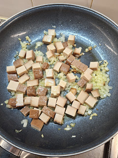 Frigideira com cubos de tofu fumado e cebola picada a saltear, alguns pedaços mais dourados e outros ainda claros, sobre um fogão.