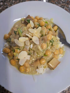 White plate with creamy tofu, chickpea, leek and mushroom curry served over white rice, topped with fresh herbs and sliced almonds.
