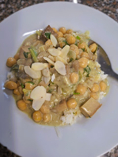 Prato branco com caril cremoso de tofu, grão, alho‑francês e cogumelos servido sobre arroz branco, decorado com ervas frescas e amêndoas laminadas.