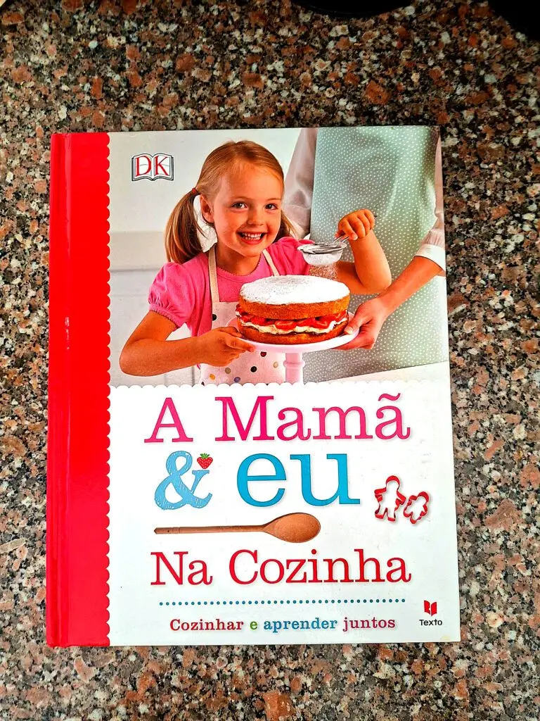Cover of the children’s book “Mom & Me in the Kitchen,” published by Texto/DK, featuring a smiling blonde girl in an apron holding a strawberry cake dusted with powdered sugar on a granite countertop.