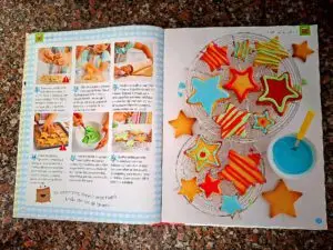 Double page spread from the book with six step-by-step photographs of the "Star Biscuits" recipe on the left, and on the right a photograph of star-shaped biscuits decorated with red, green and blue coloured icing on a cooling rack.