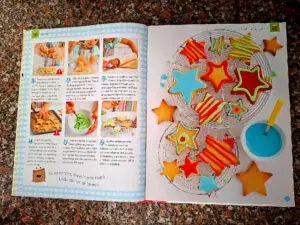 Dupla página do livro com seis fotografias do passo a passo da receita "Bolachinhas em estrela" à esquerda, e à direita uma fotografia de bolachas em forma de estrela decoradas com coberturas coloridas vermelha, verde e azul sobre uma grelha de arrefecimento.