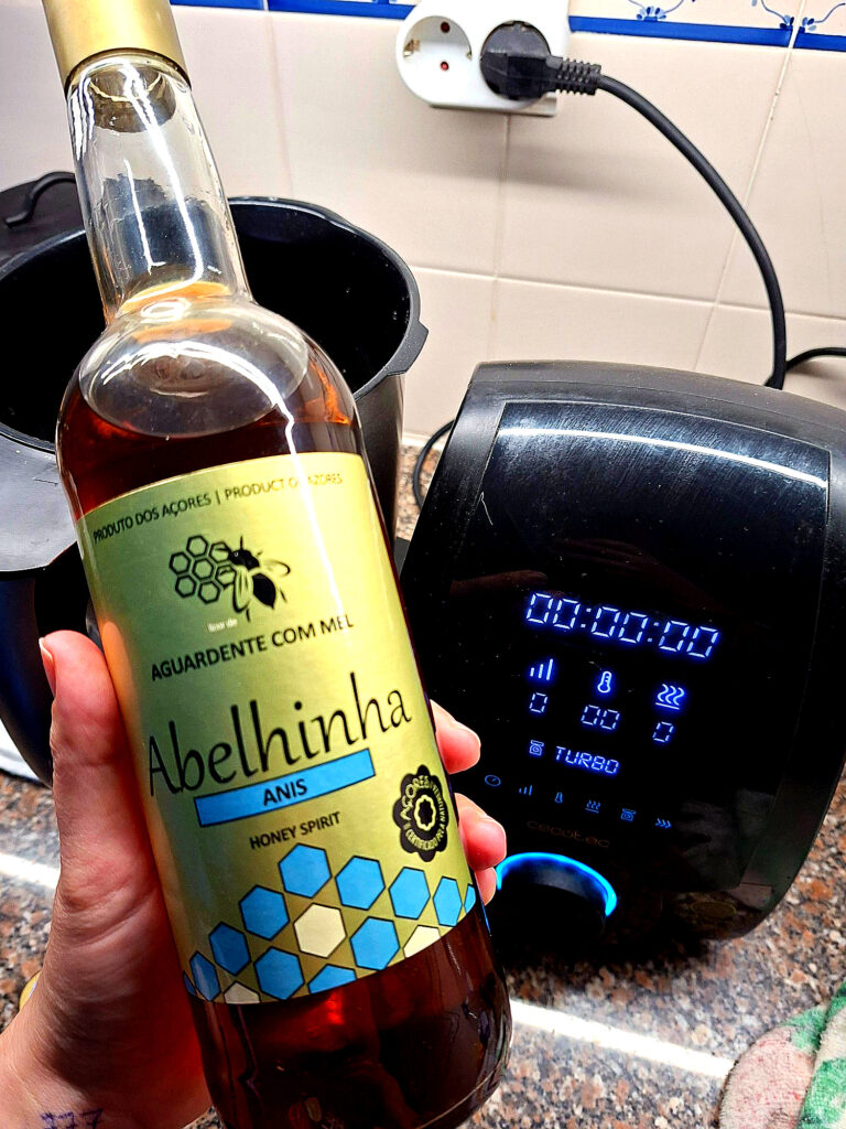 Mão a segurar uma garrafa de Aguardente Abelhinha com mel e anis, com o Robot Mambo ligado ao fundo num balcão de cozinha.