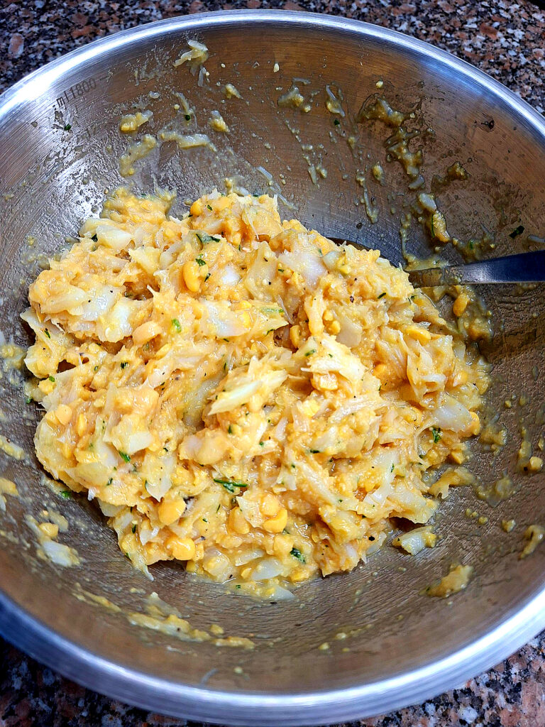 Overhead view of a stainless‑steel bowl filled with raw cod fritter batter, showing flakes of shredded cod, chickpeas, onion and green herbs, with a fork inside, on a granite countertop. Caption: When the batter looks like this — golden, fragrant and full of cod — you just know it’s going to turn out great.