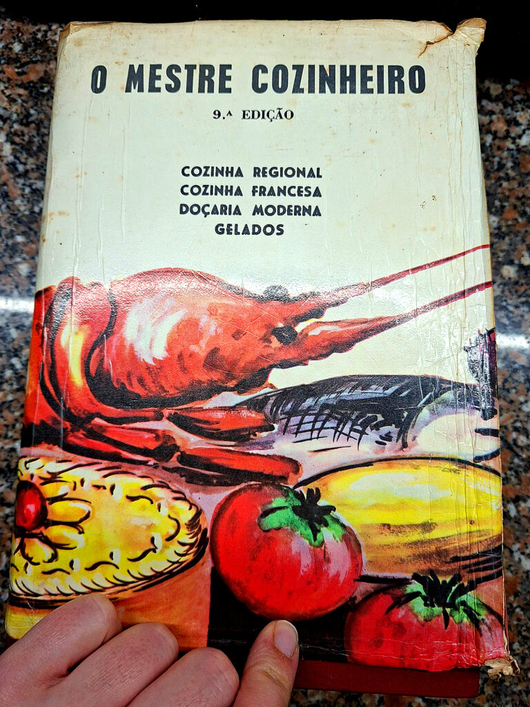 Hand holding the worn cover of O Mestre Cozinheiro, 9th Edition, featuring a vintage illustration of a lobster, tomatoes and other foods, with subtitles “Regional Cuisine, French Cuisine, Modern Pastry, Ice Creams.”