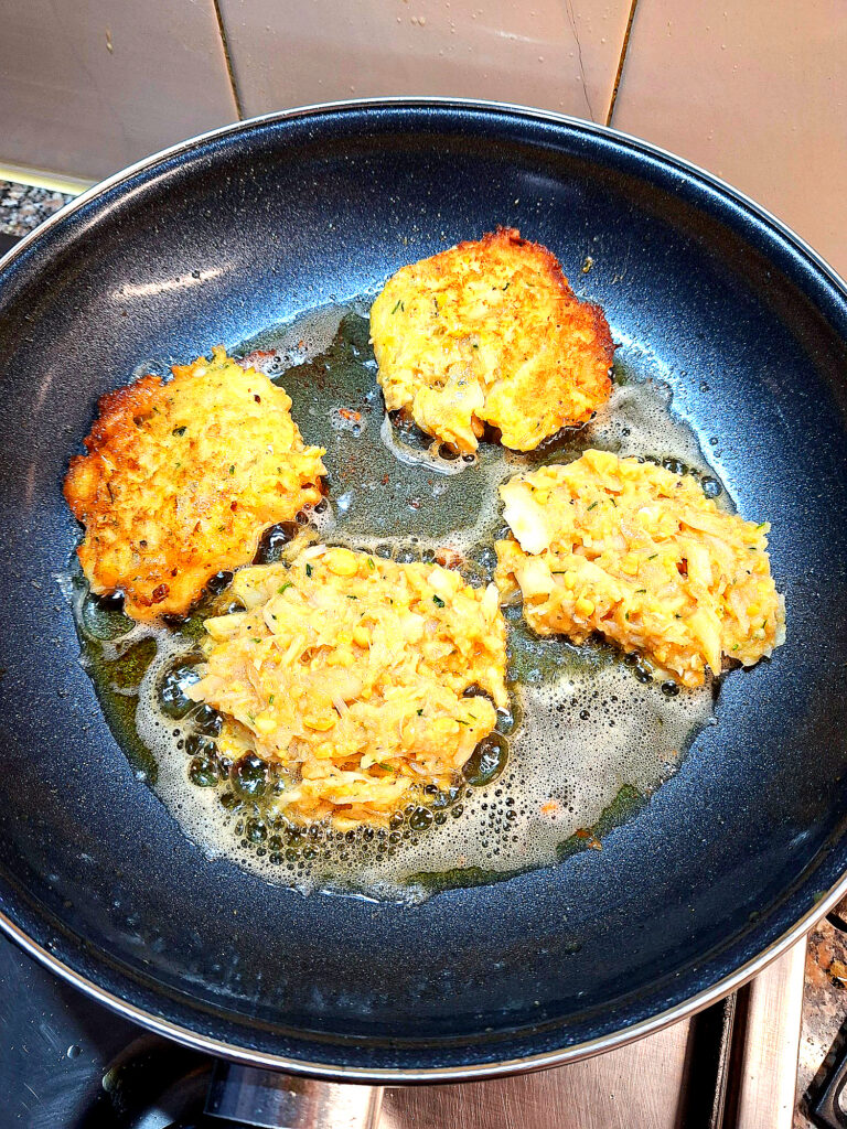 Overhead view of a dark non‑stick frying pan with four cod fritters at different frying