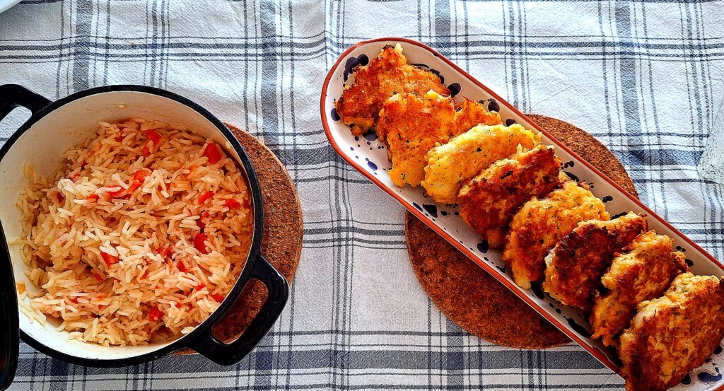 Frontal view of a blue‑and‑white checkered tablecloth with an enamelled cast‑iron pot of tomato rice on the left and a white Portuguese ceramic platter with blue dots holding golden cod fritters on the right, both on cork trivets.