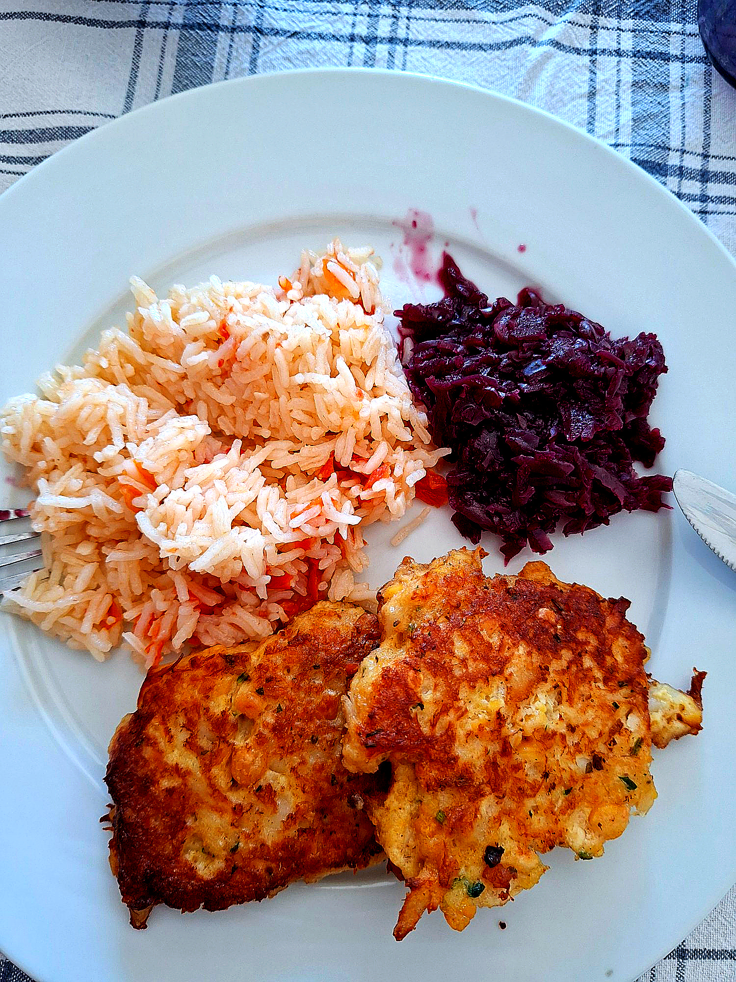 White plate with two golden cod fritters, a portion of tomato rice and cooked purple beetroot salad, on a blue‑and‑white checkered cloth with fork and knife.