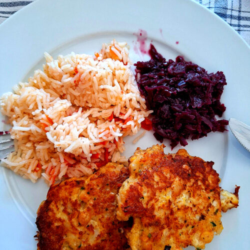 Prato branco com duas pataniscas de bacalhau douradas, porção de arroz de tomate e salada de beterraba roxa cozida, sobre toalha de xadrez azul e branca, com garfo e faca.