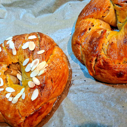 Two golden Easter braids on a baking tray, one topped with sliced almonds and the other plain.