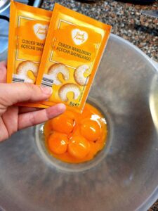 Bowl with egg yolks and a hand holding two packets of vanilla sugar over the countertop.