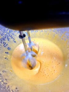 Batedeira elétrica a misturar um creme amarelo‑alaranjado numa taça, criando um movimento espiralado.