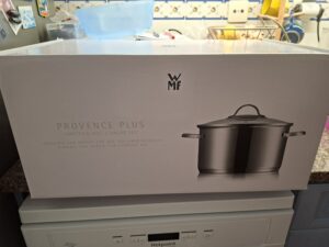 Caixa do conjunto de tachos WMF Provence Plus em aço inoxidável, com imagem do tacho e descrição em inglês e alemão, sobre máquina de lavar roupa em cozinha doméstica