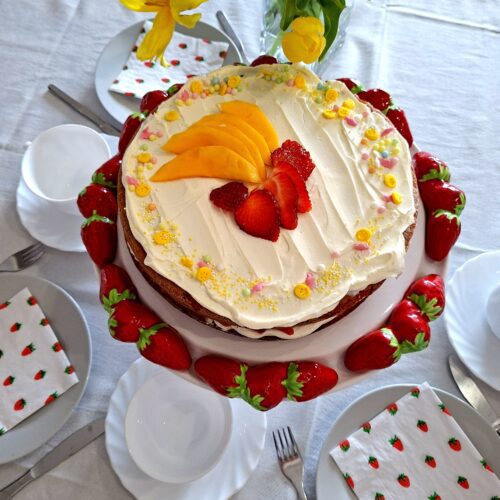 Vista aérea de bolo de limão com mascarpone e morango decorado com manga e morango numa bancada com morangos frescos, guardanapos estampados e tulipas amarelas