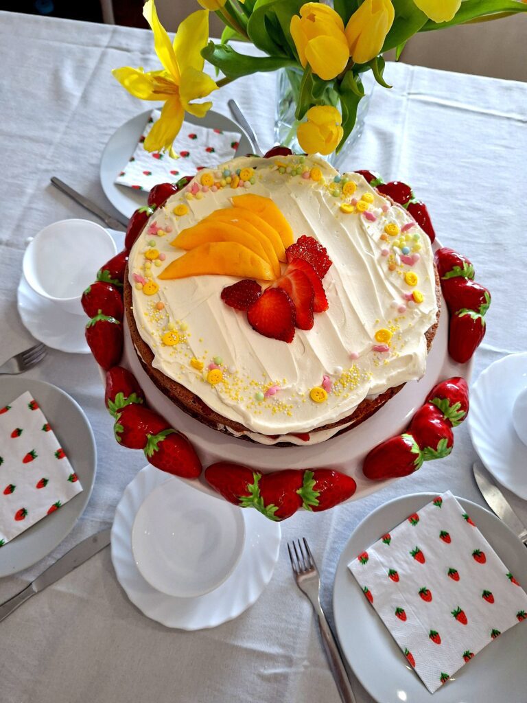 Vista aérea de bolo de limão com mascarpone e morango decorado com manga e morango numa bancada com morangos frescos, guardanapos estampados e tulipas amarelas