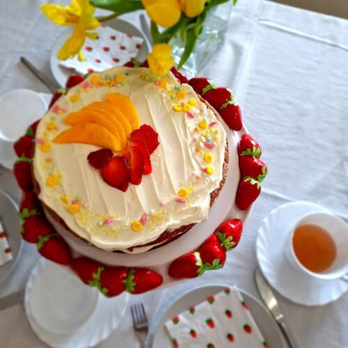 Lemon mascarpone and strawberry cake on ceramic stand with white teacup, fork and strawberry print napkin in foreground, yellow tulips in background