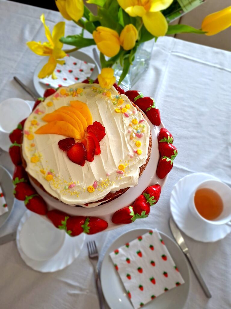 Bolo de limão com mascarpone e morango em suporte cerâmico com chávena de chá branca, garfo e guardanapo com morangos em primeiro plano, tulipas amarelas ao fundo