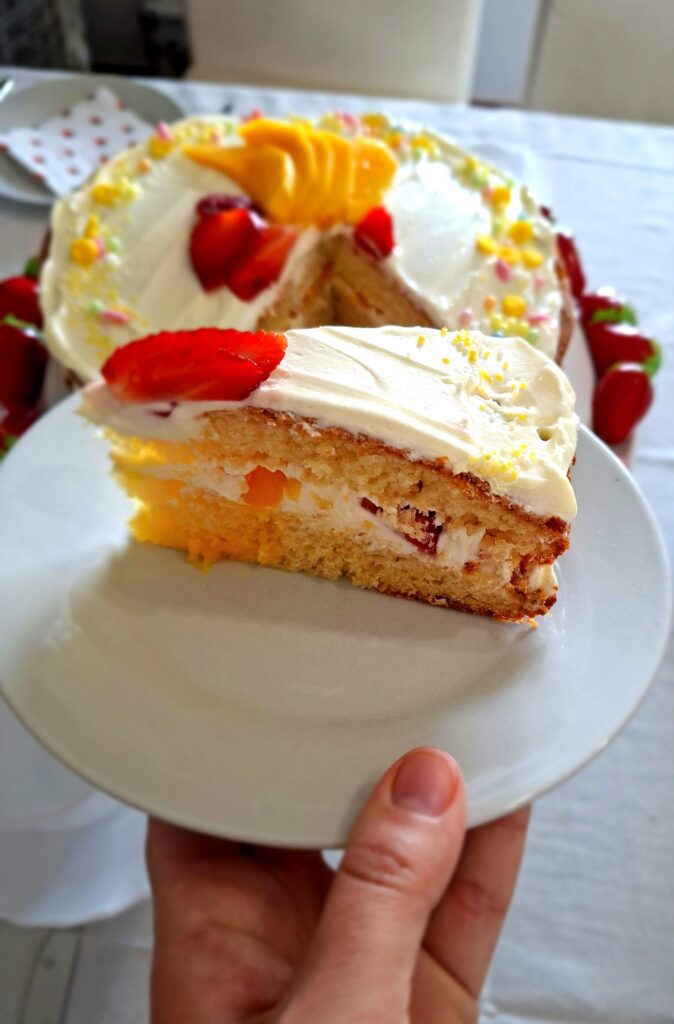 Mão a segurar prato branco com fatia de bolo de limão com recheio de mascarpone morango e pêssego, com o bolo inteiro desfocado ao fundo
