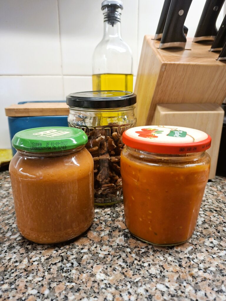 Três frascos com diferentes ingredientes — molho claro, nozes pecan e molho de tomate — dispostos numa bancada de cozinha ao lado de um bloco de facas e azeite.