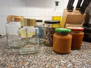 Several glass jars, some empty and others filled with pecan nuts and homemade sauce, arranged on a kitchen counter beside a knife block and olive oil.