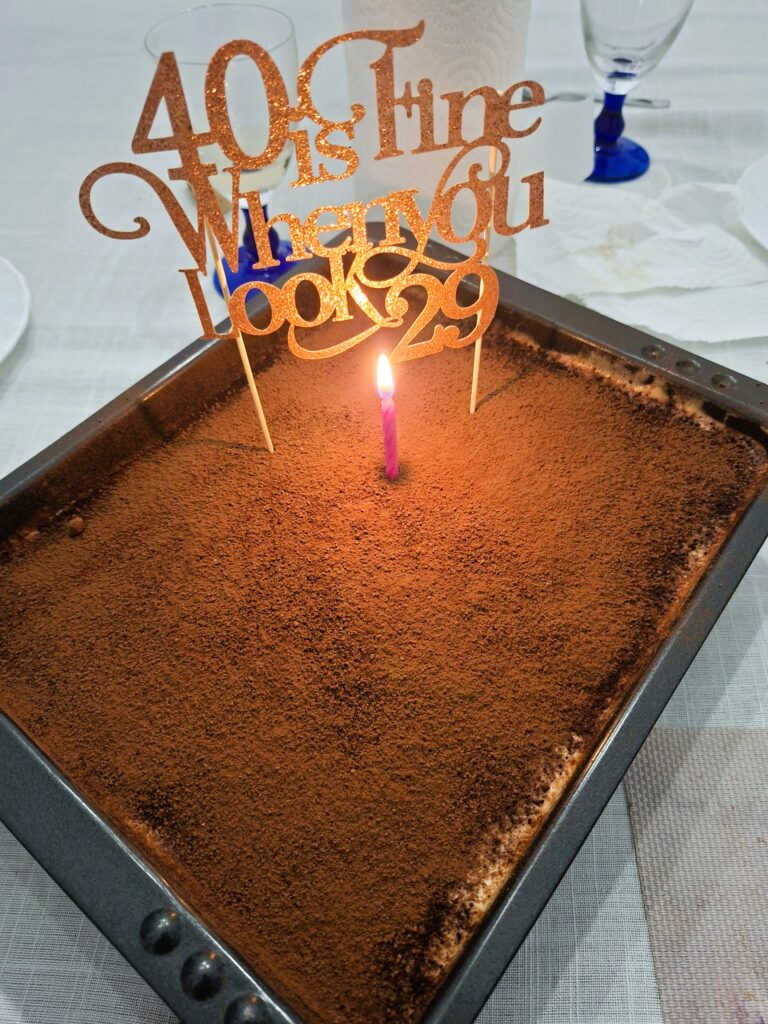  Tiramisu retangular coberto com cacau e decorado com uma vela acesa e topper dourado com mensagem de aniversário.
