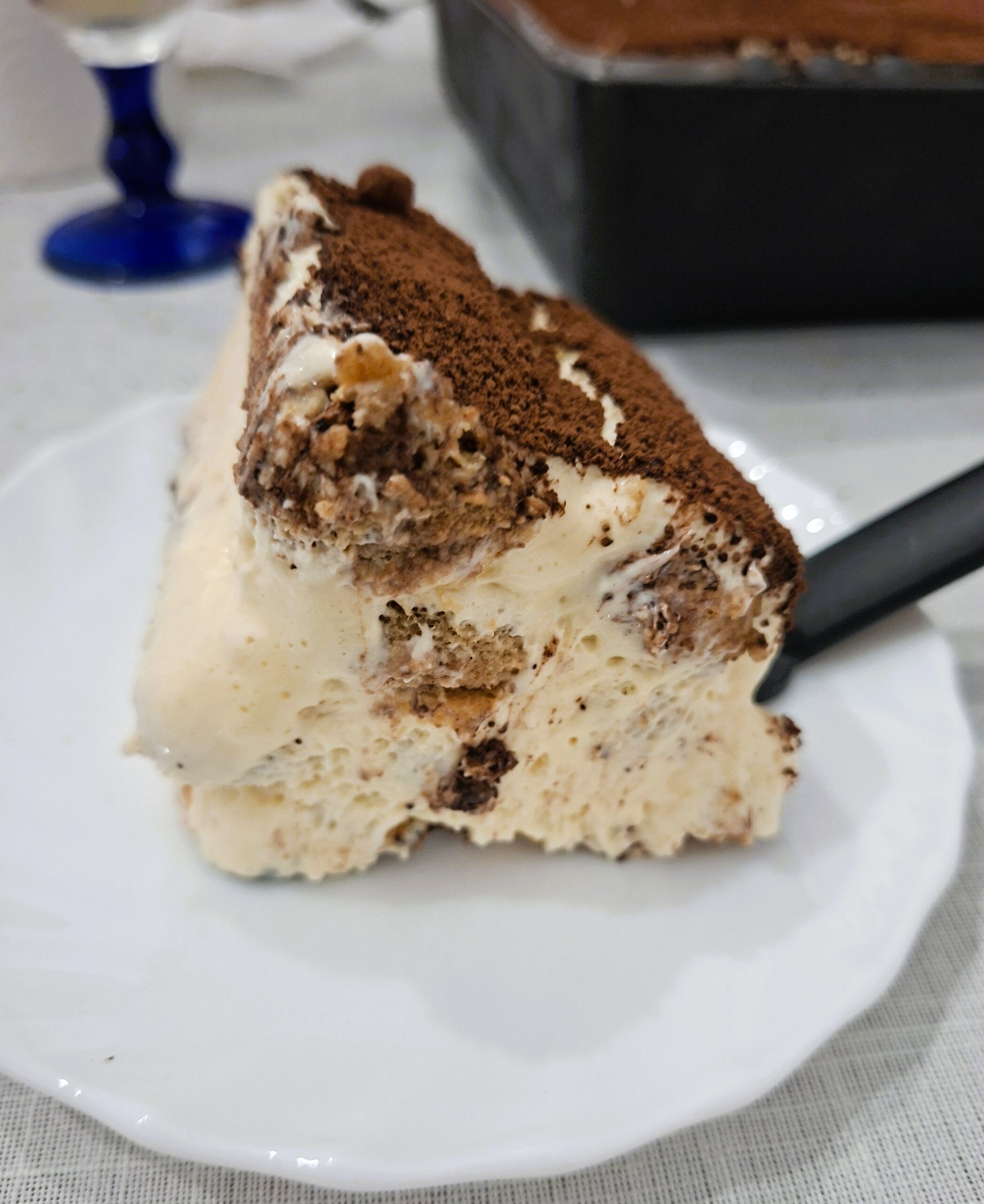 Fatia de tiramisu num prato branco, mostrando camadas de creme e biscoitos embebidos em café, polvilhada com cacau.