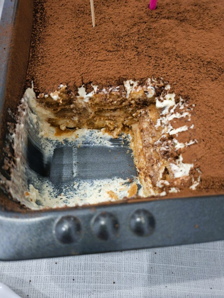 Tabuleiro de tiramisu com uma fatia retirada, revelando as camadas internas de creme e biscoitos cobertos com cacau.