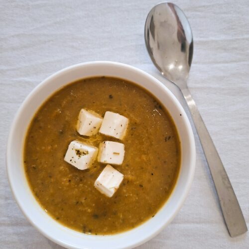 Bowl of thick brown soup topped with white cheese or tofu cubes and herbs, with a metal spoon beside it.