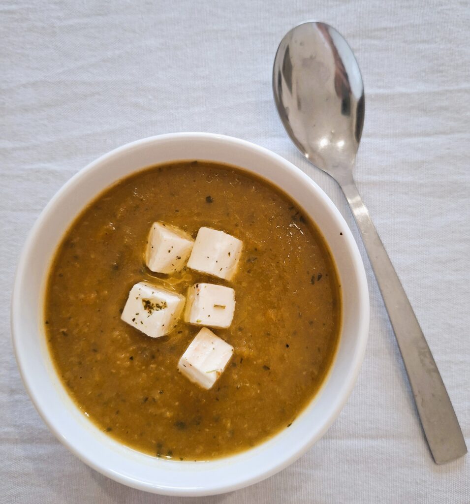 Bowl of thick brown soup topped with white cheese or tofu cubes and herbs, with a metal spoon beside it.