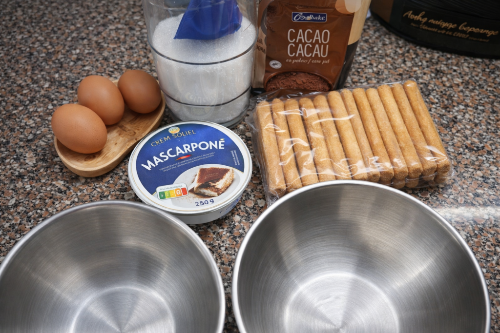 Ingredientes para tiramisu dispostos numa bancada, incluindo ovos, mascarpone, açúcar, cacau em pó e palitos de champanhe.