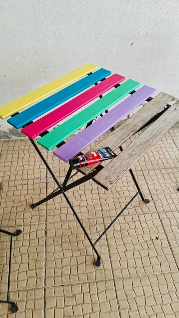 Wooden table partially painted in bright colors, with a tube of acrylic paint resting on the unpainted slats.
