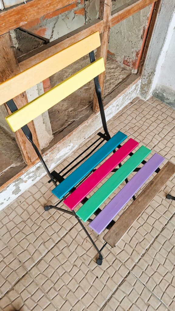 Wooden chair with brightly painted slats placed in a space with an unfinished wall.