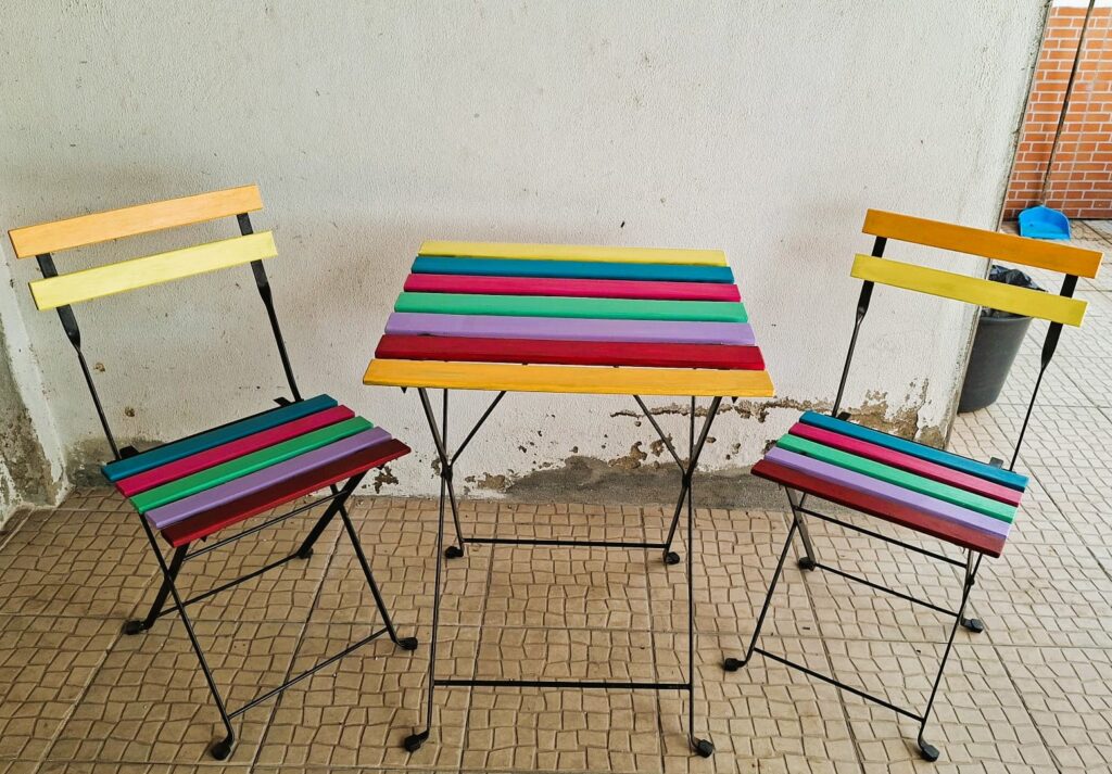 Mesa e duas cadeiras com ripas de madeira pintadas em cores vivas, colocadas num espaço exterior simples.