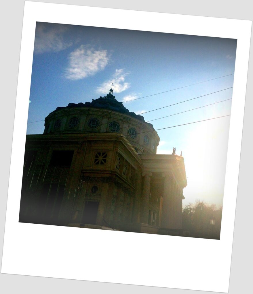 Tilted polaroid-style photograph of the Romanian Athenaeum in Bucharest seen in backlight at dusk, with the dome and neoclassical facade in golden silhouette against a blue sky with clouds and visible electric wires.