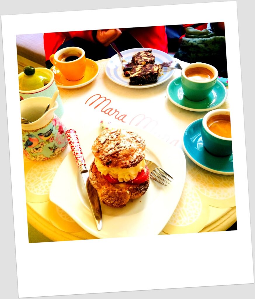 Table at Mara Mura patisserie with a choux bun filled with cream and strawberries on a white plate with red fork and knife, two espressos in turquoise and orange cups, a chocolate brownie in the background, a floral sugar bowl and napkins, with the Mara Mura branded paper tablecloth visible.